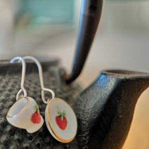 tiny tea cup and saucer earrings in porcelain with a red strawberry pattern hang off a metal tea pot